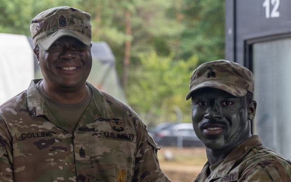 Two U.S. Army soldiers in OCP (Operational Camouflage Pattern) uniforms are posing together while shaking hands and exchanging what appears to be a certificate or document. The soldier on the left, identified by his name tape as 'Collins,' wears a Command Sergeant Major rank insignia on his cap, suggesting this is an award or recognition ceremony. The soldier on the right has tactical face paint applied, indicating recent or ongoing field training exercises.