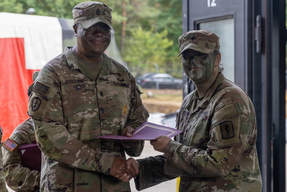 Two U.S. Army soldiers in MultiCam/OCP camouflage uniforms are shaking hands while jointly holding a purple folder or certificate, suggesting an award or recognition ceremony. The soldier on the left has the name tape 'COLLINS' and visible rank insignia of a Senior NCO (Sergeant Major or Command Sergeant Major) on their patrol cap. The soldier on the right has green camouflage face paint applied, indicating recent field exercise participation.