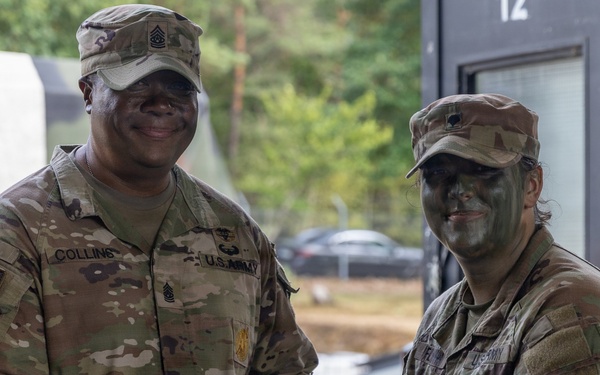 Two U.S. Army soldiers in MultiCam/OCP camouflage uniforms are shaking hands while jointly holding a purple folder or certificate, suggesting an award or recognition ceremony. The soldier on the left has the name tape 'COLLINS' and visible rank insignia of a Senior NCO (Sergeant Major or Command Sergeant Major) on their patrol cap. The soldier on the right has green camouflage face paint applied, indicating recent field exercise participation.