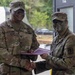 Two U.S. Army soldiers in MultiCam/OCP camouflage uniforms are shaking hands while jointly holding a purple folder or certificate, suggesting an award or recognition ceremony. The soldier on the left has the name tape 'COLLINS' and visible rank insignia of a Senior NCO (Sergeant Major or Command Sergeant Major) on their patrol cap. The soldier on the right has green camouflage face paint applied, indicating recent field exercise participation.