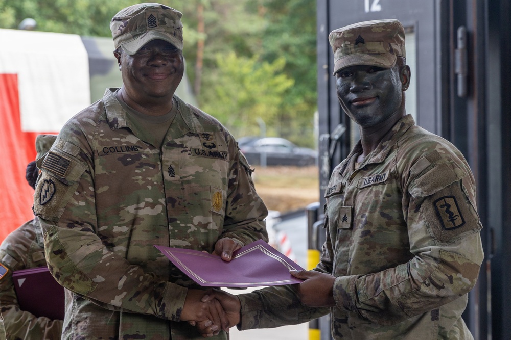 Two U.S. Army soldiers are posing together while jointly holding a purple certificate or award folder. The soldier on the left, identified by name tape as 'Collins,' appears to be a senior NCO presenting an award or commendation to the soldier on the right, who is wearing tactical face paint. Both are in OCP (Operational Camouflage Pattern) uniforms.