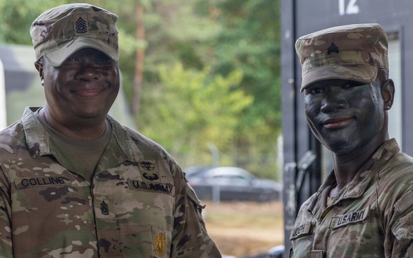 Two U.S. Army soldiers are posing together while jointly holding a purple certificate or award folder. The soldier on the left, identified by name tape as 'Collins,' appears to be a senior NCO presenting an award or commendation to the soldier on the right, who is wearing tactical face paint. Both are in OCP (Operational Camouflage Pattern) uniforms.