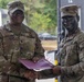 Two U.S. Army soldiers are posing together while jointly holding a purple certificate or award folder. The soldier on the left, identified by name tape as 'Collins,' appears to be a senior NCO presenting an award or commendation to the soldier on the right, who is wearing tactical face paint. Both are in OCP (Operational Camouflage Pattern) uniforms.