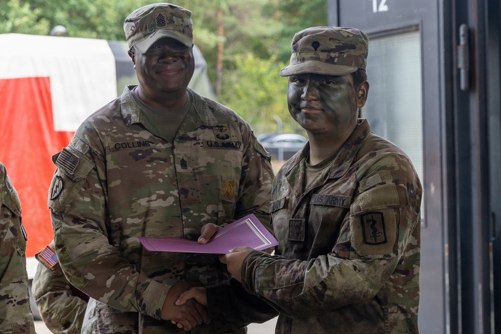 Two U.S. Army soldiers in MultiCam (OCP) uniforms are shaking hands while jointly holding a purple folder or certificate, suggesting an award or recognition ceremony. The soldier on the left is identified by name tape as 'Collins' and wears Senior NCO rank insignia (First Sergeant or Sergeant Major chevrons on collar). The soldier on the right has tactical face camouflage paint applied and wears a medical corps branch insignia patch on their right shoulder.