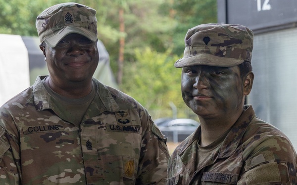 Two U.S. Army soldiers in MultiCam (OCP) uniforms are shaking hands while jointly holding a purple folder or certificate, suggesting an award or recognition ceremony. The soldier on the left is identified by name tape as 'Collins' and wears Senior NCO rank insignia (First Sergeant or Sergeant Major chevrons on collar). The soldier on the right has tactical face camouflage paint applied and wears a medical corps branch insignia patch on their right shoulder.
