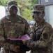 Two U.S. Army soldiers in MultiCam (OCP) uniforms are shaking hands while jointly holding a purple folder or certificate, suggesting an award or recognition ceremony. The soldier on the left is identified by name tape as 'Collins' and wears Senior NCO rank insignia (First Sergeant or Sergeant Major chevrons on collar). The soldier on the right has tactical face camouflage paint applied and wears a medical corps branch insignia patch on their right shoulder.