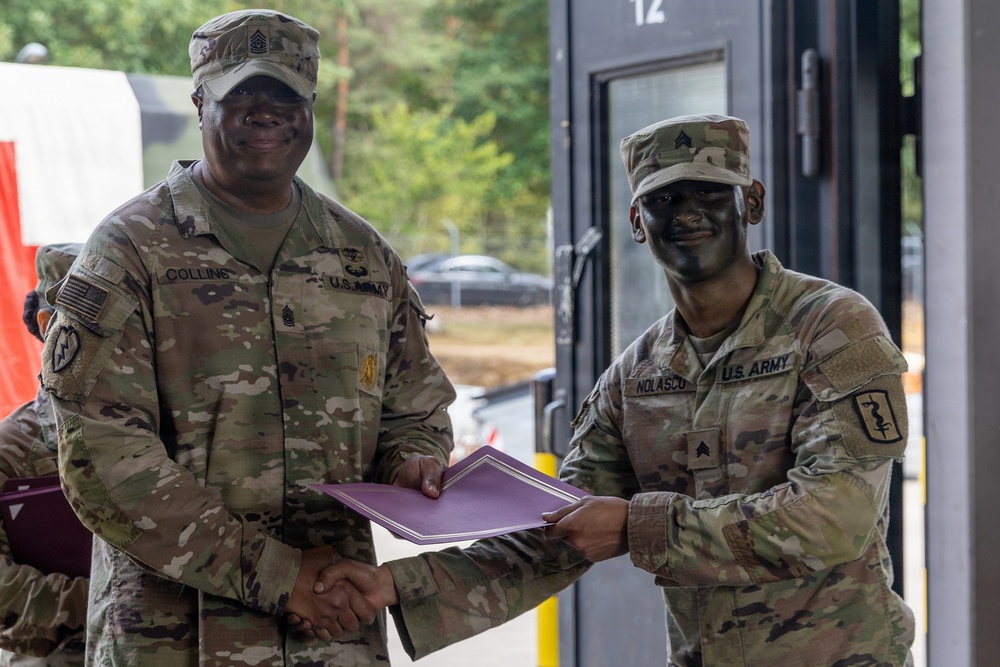 Two U.S. Army soldiers in MultiCam (OCP) uniforms are shaking hands while jointly holding a purple folder or certificate, suggesting an award or recognition ceremony. The soldier on the left, identified by nametape as 'Collins,' holds a senior NCO rank (Command Sergeant Major based on collar insignia), while the soldier on the right, identified as 'Nolasco,' holds a Staff Sergeant rank. Both soldiers are smiling and wearing combat face paint, indicating a field training environment.