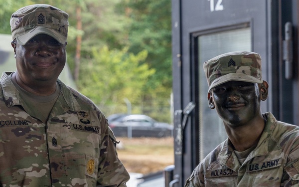 Two U.S. Army soldiers in MultiCam (OCP) uniforms are shaking hands while jointly holding a purple folder or certificate, suggesting an award or recognition ceremony. The soldier on the left, identified by nametape as 'Collins,' holds a senior NCO rank (Command Sergeant Major based on collar insignia), while the soldier on the right, identified as 'Nolasco,' holds a Staff Sergeant rank. Both soldiers are smiling and wearing combat face paint, indicating a field training environment.