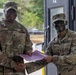 Two U.S. Army soldiers in MultiCam (OCP) uniforms are shaking hands while jointly holding a purple folder or certificate, suggesting an award or recognition ceremony. The soldier on the left, identified by nametape as 'Collins,' holds a senior NCO rank (Command Sergeant Major based on collar insignia), while the soldier on the right, identified as 'Nolasco,' holds a Staff Sergeant rank. Both soldiers are smiling and wearing combat face paint, indicating a field training environment.