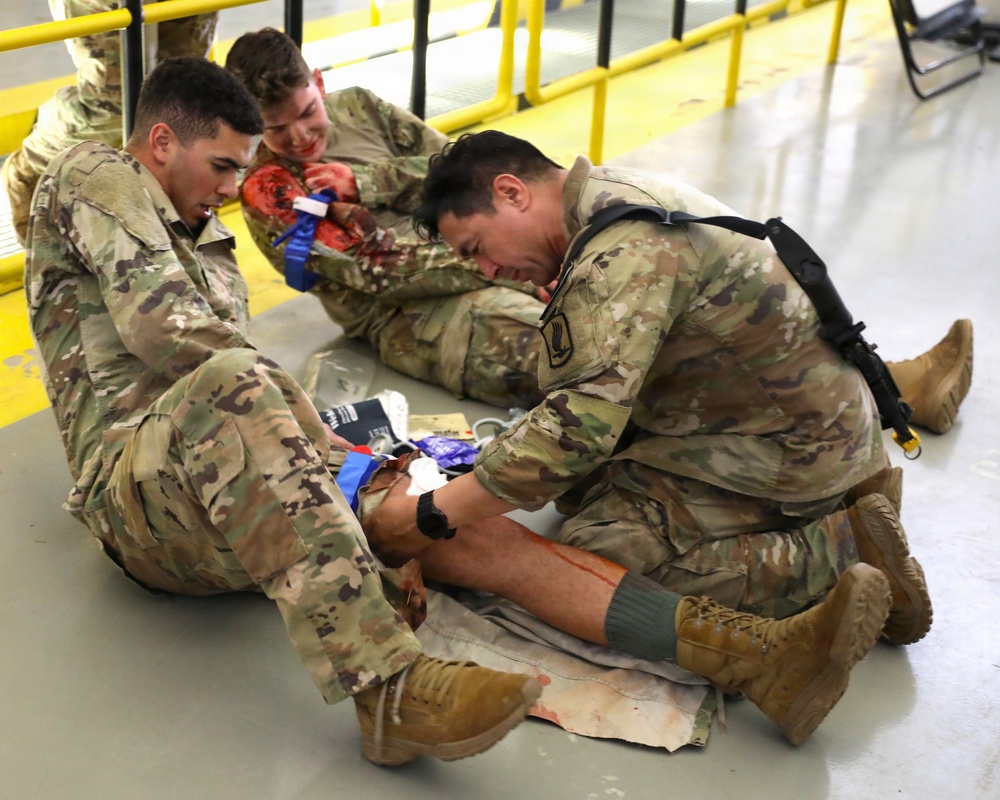 Military personnel in OCP (Operational Camouflage Pattern) uniforms are conducting a combat casualty care training exercise on an indoor floor. Two soldiers are treating a simulated casualty who has moulage (simulated wounds with fake blood) applied to their arm and leg. Medical supplies including bandages and packaging are visible on the floor around them.