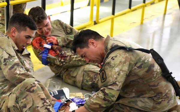 Military personnel in OCP (Operational Camouflage Pattern) uniforms are conducting a combat casualty care training exercise on an indoor floor. Two soldiers are treating a simulated casualty who has moulage (simulated wounds with fake blood) applied to their arm and leg. Medical supplies including bandages and packaging are visible on the floor around them.