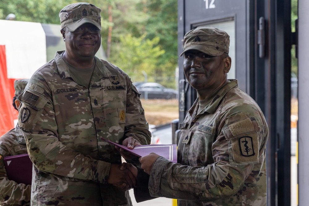 Two U.S. Army soldiers in MultiCam (OCP) uniforms are shaking hands while jointly holding a purple folder or certificate, suggesting an award or recognition ceremony. The soldier on the left has a name tape reading 'COLLINS' and displays senior NCO rank insignia, while the soldier on the right wears a unit patch with a medical caduceus symbol. The setting appears to be an outdoor military installation or training area.