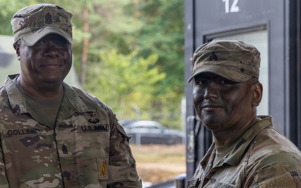 Two U.S. Army soldiers in MultiCam (OCP) uniforms are shaking hands while jointly holding a purple folder or certificate, suggesting an award or recognition ceremony. The soldier on the left has a name tape reading 'COLLINS' and displays senior NCO rank insignia, while the soldier on the right wears a unit patch with a medical caduceus symbol. The setting appears to be an outdoor military installation or training area.