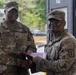 Two U.S. Army soldiers in MultiCam (OCP) uniforms are shaking hands while jointly holding a purple folder or certificate, suggesting an award or recognition ceremony. The soldier on the left has a name tape reading 'COLLINS' and displays senior NCO rank insignia, while the soldier on the right wears a unit patch with a medical caduceus symbol. The setting appears to be an outdoor military installation or training area.