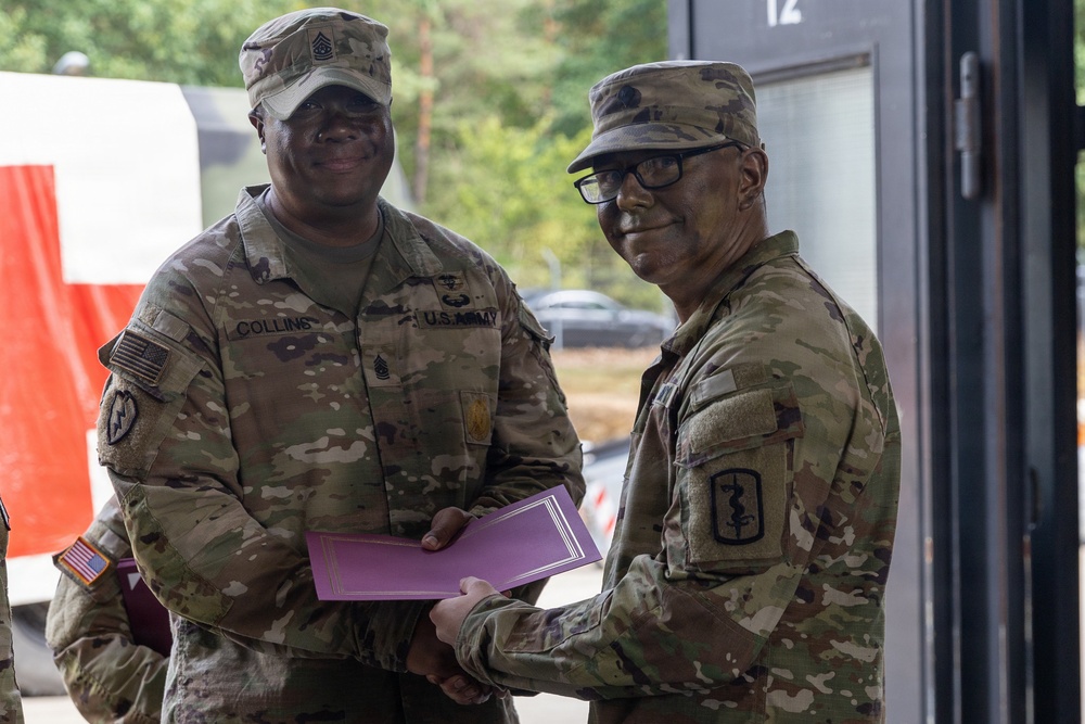 Two U.S. Army soldiers in MultiCam/OCP camouflage uniforms are posing together while jointly holding a purple folder or certificate, suggesting an award or recognition ceremony. The soldier on the left has the name tape 'COLLINS' visible and wears Senior NCO rank insignia on his cap. Both soldiers are smiling, indicating a positive ceremonial occasion.
