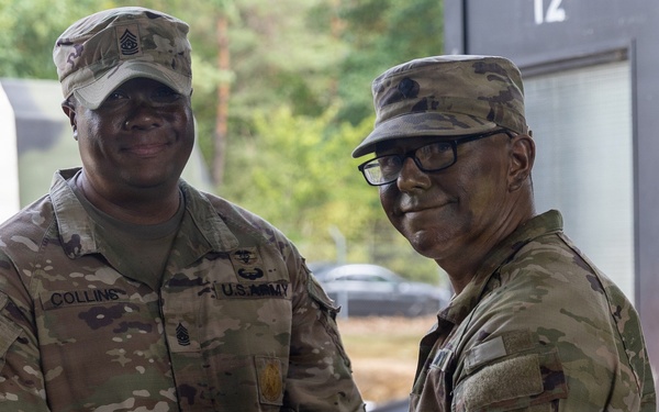 Two U.S. Army soldiers in MultiCam/OCP camouflage uniforms are posing together while jointly holding a purple folder or certificate, suggesting an award or recognition ceremony. The soldier on the left has the name tape 'COLLINS' visible and wears Senior NCO rank insignia on his cap. Both soldiers are smiling, indicating a positive ceremonial occasion.