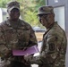 Two U.S. Army soldiers in MultiCam/OCP camouflage uniforms are posing together while jointly holding a purple folder or certificate, suggesting an award or recognition ceremony. The soldier on the left has the name tape 'COLLINS' visible and wears Senior NCO rank insignia on his cap. Both soldiers are smiling, indicating a positive ceremonial occasion.