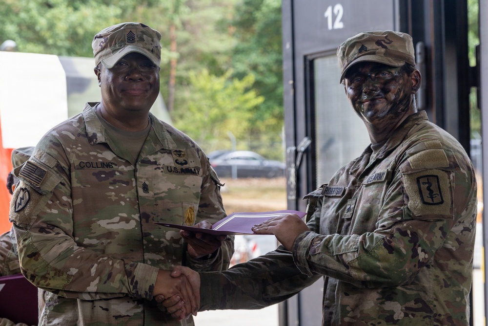 Two U.S. Army soldiers are shaking hands while jointly holding what appears to be a certificate or award document in a military setting. The soldier on the left, identified by name tape as 'Collins,' wears a Sergeant Major rank insignia and is smiling. The soldier on the right, identified as 'Robertson,' has camouflage face paint applied and wears a Medical Corps branch insignia patch.