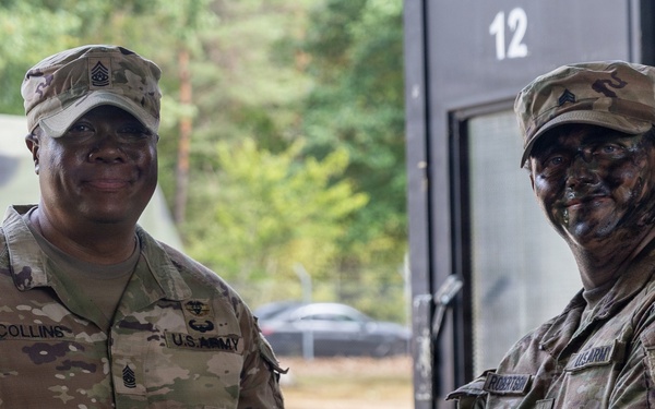 Two U.S. Army soldiers are shaking hands while jointly holding what appears to be a certificate or award document in a military setting. The soldier on the left, identified by name tape as 'Collins,' wears a Sergeant Major rank insignia and is smiling. The soldier on the right, identified as 'Robertson,' has camouflage face paint applied and wears a Medical Corps branch insignia patch.