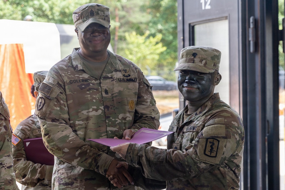 Two U.S. Army soldiers are engaged in what appears to be an award or certificate presentation ceremony. The senior soldier, identified by nametape as 'Collins,' is presenting a document in a purple folder to a junior soldier who has tactical face camouflage paint applied. The setting appears to be an outdoor or semi-outdoor military installation.