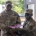 Two U.S. Army soldiers are engaged in what appears to be an award or certificate presentation ceremony. The senior soldier, identified by nametape as 'Collins,' is presenting a document in a purple folder to a junior soldier who has tactical face camouflage paint applied. The setting appears to be an outdoor or semi-outdoor military installation.