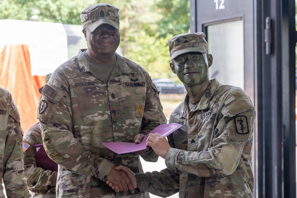 Two U.S. Army soldiers are shaking hands while jointly holding a pink/purple folder or certificate, suggesting an award or recognition ceremony. The soldier on the left, identified by his name tape as 'Collins,' wears Senior NCO rank insignia, while the soldier on the right has green camouflage face paint applied and wears a medical branch insignia. The setting appears to be an outdoor military installation or training area.