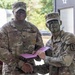 Two U.S. Army soldiers are shaking hands while jointly holding a pink/purple folder or certificate, suggesting an award or recognition ceremony. The soldier on the left, identified by his name tape as 'Collins,' wears Senior NCO rank insignia, while the soldier on the right has green camouflage face paint applied and wears a medical branch insignia. The setting appears to be an outdoor military installation or training area.