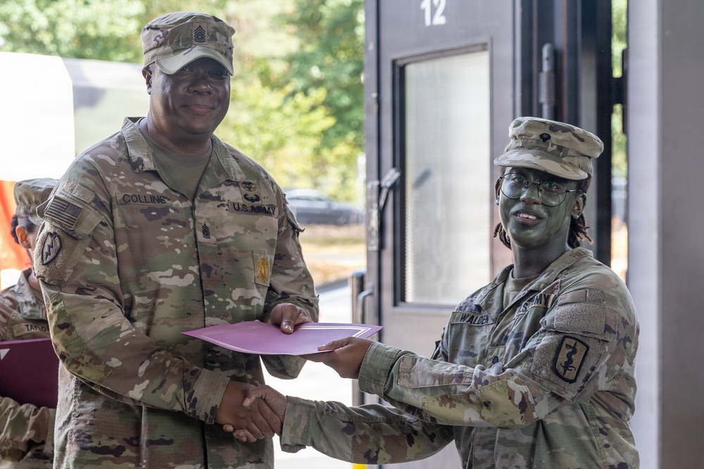 Two U.S. Army soldiers in OCP (Operational Camouflage Pattern) uniforms are shaking hands while exchanging a document in a pink/purple folder, suggesting a formal recognition or award ceremony. The soldier on the left, identified by name tape as 'Collins,' appears to be a senior NCO based on rank insignia visible on the cap. A third soldier is partially visible in the background.