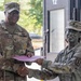 Two U.S. Army soldiers in OCP (Operational Camouflage Pattern) uniforms are shaking hands while exchanging a document in a pink/purple folder, suggesting a formal recognition or award ceremony. The soldier on the left, identified by name tape as 'Collins,' appears to be a senior NCO based on rank insignia visible on the cap. A third soldier is partially visible in the background.