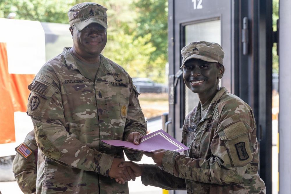 Two U.S. Army soldiers in OCP (Operational Camouflage Pattern) uniforms are shaking hands while one presents a certificate or award folder to the other. The soldier on the left, identified by name tape as 'Collins,' appears to be a senior NCO based on rank insignia visible on the cap. The ceremony appears to be taking place outdoors near a building entrance marked with the number 12.