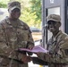 Two U.S. Army soldiers in OCP (Operational Camouflage Pattern) uniforms are shaking hands while one presents a certificate or award folder to the other. The soldier on the left, identified by name tape as 'Collins,' appears to be a senior NCO based on rank insignia visible on the cap. The ceremony appears to be taking place outdoors near a building entrance marked with the number 12.