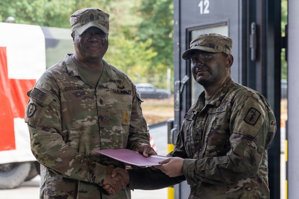 Two U.S. Army soldiers in OCP (Operational Camouflage Pattern) uniforms are shaking hands while jointly holding a maroon/burgundy folder or certificate, suggesting an award or recognition ceremony. The soldier on the left, identified by his name tape as 'COLLINS,' wears a Command Sergeant Major rank insignia on his cap, indicating a senior NCO presenting an award. The ceremony appears to be taking place outdoors near a building with the number '12' visible.