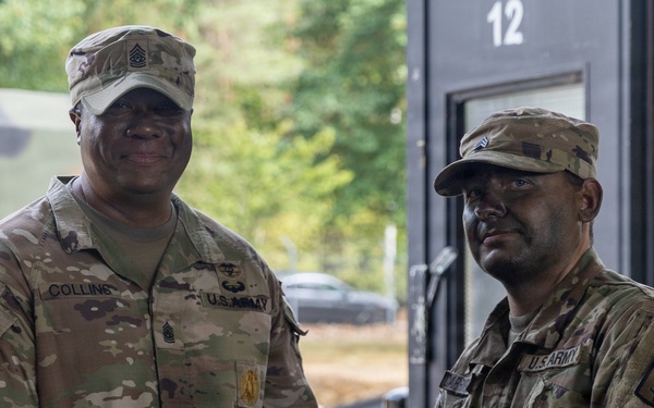 Two U.S. Army soldiers in OCP (Operational Camouflage Pattern) uniforms are shaking hands while jointly holding a maroon/burgundy folder or certificate, suggesting an award or recognition ceremony. The soldier on the left, identified by his name tape as 'COLLINS,' wears a Command Sergeant Major rank insignia on his cap, indicating a senior NCO presenting an award. The ceremony appears to be taking place outdoors near a building with the number '12' visible.