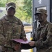 Two U.S. Army soldiers in OCP (Operational Camouflage Pattern) uniforms are shaking hands while jointly holding a maroon/burgundy folder or certificate, suggesting an award or recognition ceremony. The soldier on the left, identified by his name tape as 'COLLINS,' wears a Command Sergeant Major rank insignia on his cap, indicating a senior NCO presenting an award. The ceremony appears to be taking place outdoors near a building with the number '12' visible.