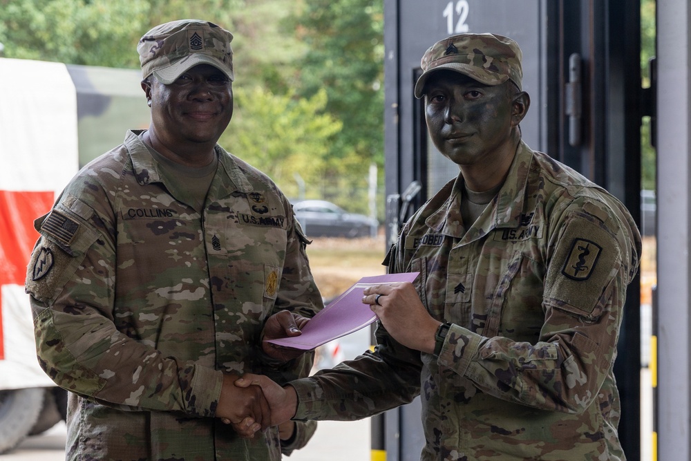 Two U.S. Army soldiers are shaking hands in what appears to be an award or recognition ceremony. The soldier on the left, identified by nametape as 'COLLINS,' holds a senior NCO rank and is presenting a document in a pink envelope to the soldier on the right, whose nametape reads 'OBEDO' (partially visible), who is wearing tactical face camouflage paint. Both are in OCP (Operational Camouflage Pattern) uniforms.