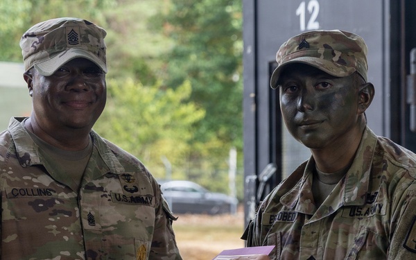 Two U.S. Army soldiers are shaking hands in what appears to be an award or recognition ceremony. The soldier on the left, identified by nametape as 'COLLINS,' holds a senior NCO rank and is presenting a document in a pink envelope to the soldier on the right, whose nametape reads 'OBEDO' (partially visible), who is wearing tactical face camouflage paint. Both are in OCP (Operational Camouflage Pattern) uniforms.
