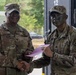 Two U.S. Army soldiers are shaking hands in what appears to be an award or recognition ceremony. The soldier on the left, identified by nametape as 'COLLINS,' holds a senior NCO rank and is presenting a document in a pink envelope to the soldier on the right, whose nametape reads 'OBEDO' (partially visible), who is wearing tactical face camouflage paint. Both are in OCP (Operational Camouflage Pattern) uniforms.