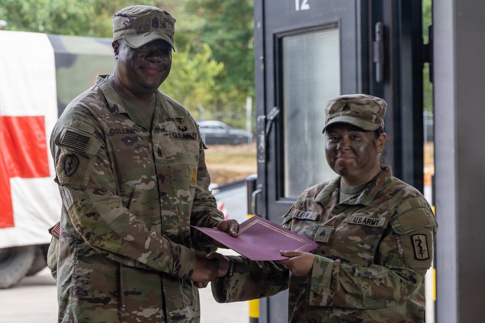 Two U.S. Army soldiers in OCP (Operational Camouflage Pattern) uniforms are engaged in what appears to be an award or certificate presentation ceremony. The taller soldier on the left, identified by nametape as 'Collins,' is presenting a maroon/burgundy folder to the soldier on the right, identified as 'LEI.' Both soldiers are smiling, suggesting a positive recognition event.