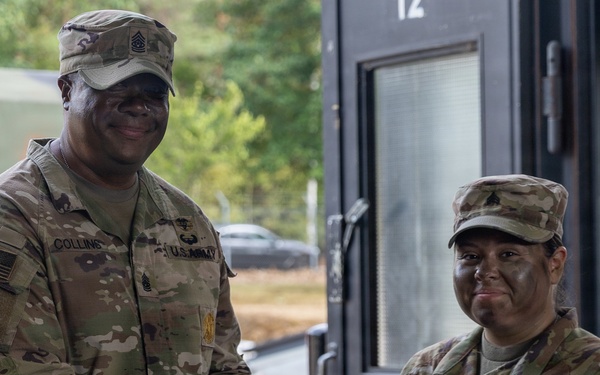 Two U.S. Army soldiers in OCP (Operational Camouflage Pattern) uniforms are engaged in what appears to be an award or certificate presentation ceremony. The taller soldier on the left, identified by nametape as 'Collins,' is presenting a maroon/burgundy folder to the soldier on the right, identified as 'LEI.' Both soldiers are smiling, suggesting a positive recognition event.