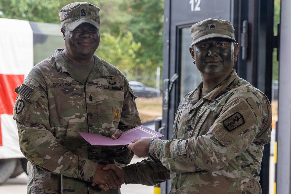 Two U.S. Army soldiers in OCP (Operational Camouflage Pattern) uniforms are shaking hands while jointly holding a certificate or award folder. The soldier on the left, identified by name tape as 'Collins,' appears to be a senior NCO presenting an award or recognition to the soldier on the right. Both soldiers have tactical face paint applied, suggesting this may be occurring during or after a field training exercise.