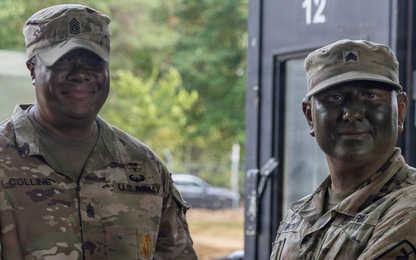 Two U.S. Army soldiers in OCP (Operational Camouflage Pattern) uniforms are shaking hands while jointly holding a certificate or award folder. The soldier on the left, identified by name tape as 'Collins,' appears to be a senior NCO presenting an award or recognition to the soldier on the right. Both soldiers have tactical face paint applied, suggesting this may be occurring during or after a field training exercise.