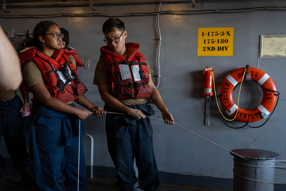 Two naval personnel wearing red life vests are handling a rope/line aboard what appears to be the USS Abraham Lincoln (CVN-72). They are engaged in line-handling training or a mooring exercise on the ship's deck. The setting is an interior deck area with safety equipment and divisional markings visible on the bulkhead.