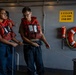 Two naval personnel wearing red life vests are handling a rope/line aboard what appears to be the USS Abraham Lincoln (CVN-72). They are engaged in line-handling training or a mooring exercise on the ship's deck. The setting is an interior deck area with safety equipment and divisional markings visible on the bulkhead.
