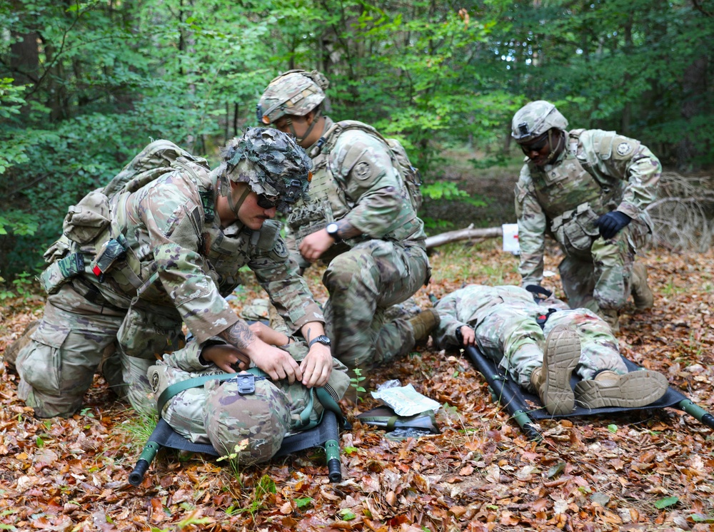 Military personnel in MultiCam/OCP camouflage uniforms are conducting a tactical combat casualty care (TCCC) training exercise in a wooded area. Four soldiers are attending to two simulated casualties lying on litters/stretchers on a leaf-covered forest floor. The scene appears to be a field training exercise (FTX) based on the controlled, instructional nature of the activity.
