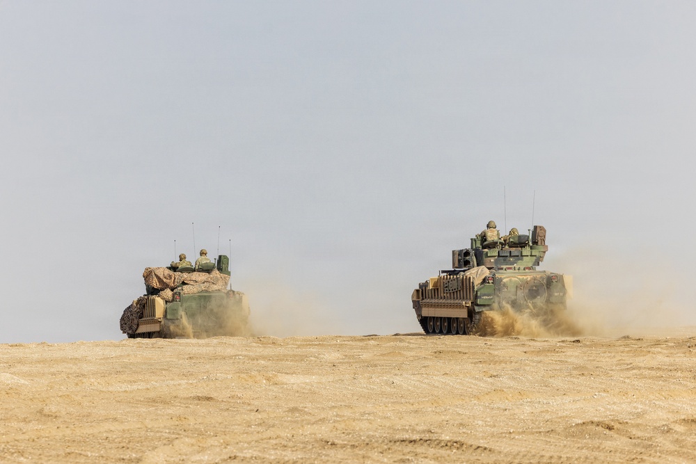 Two military armored fighting vehicles are traversing a flat, arid desert terrain, kicking up dust clouds as they move. Both vehicles carry personnel visible in open hatches on top. The vehicles appear to be in a convoy or formation during what appears to be a military exercise or operation.