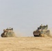 Two military armored fighting vehicles are traversing a flat, arid desert terrain, kicking up dust clouds as they move. Both vehicles carry personnel visible in open hatches on top. The vehicles appear to be in a convoy or formation during what appears to be a military exercise or operation.