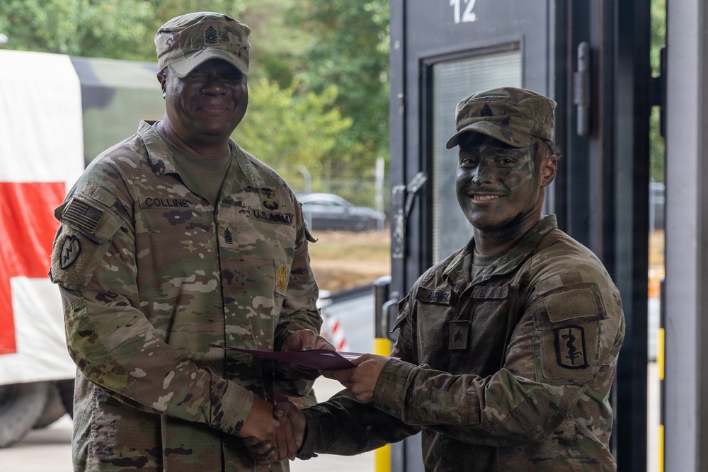 Two U.S. Army soldiers are shaking hands while exchanging what appears to be a certificate or document, likely during an award or recognition ceremony. The senior soldier on the left, identified by the name tape 'COLLINS,' wears senior NCO rank insignia, while the soldier on the right has camouflage face paint applied. The setting appears to be an outdoor or semi-covered military facility.