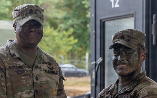 Two U.S. Army soldiers are shaking hands while exchanging what appears to be a certificate or document, likely during an award or recognition ceremony. The senior soldier on the left, identified by the name tape 'COLLINS,' wears senior NCO rank insignia, while the soldier on the right has camouflage face paint applied. The setting appears to be an outdoor or semi-covered military facility.