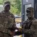 Two U.S. Army soldiers are shaking hands while exchanging what appears to be a certificate or document, likely during an award or recognition ceremony. The senior soldier on the left, identified by the name tape 'COLLINS,' wears senior NCO rank insignia, while the soldier on the right has camouflage face paint applied. The setting appears to be an outdoor or semi-covered military facility.