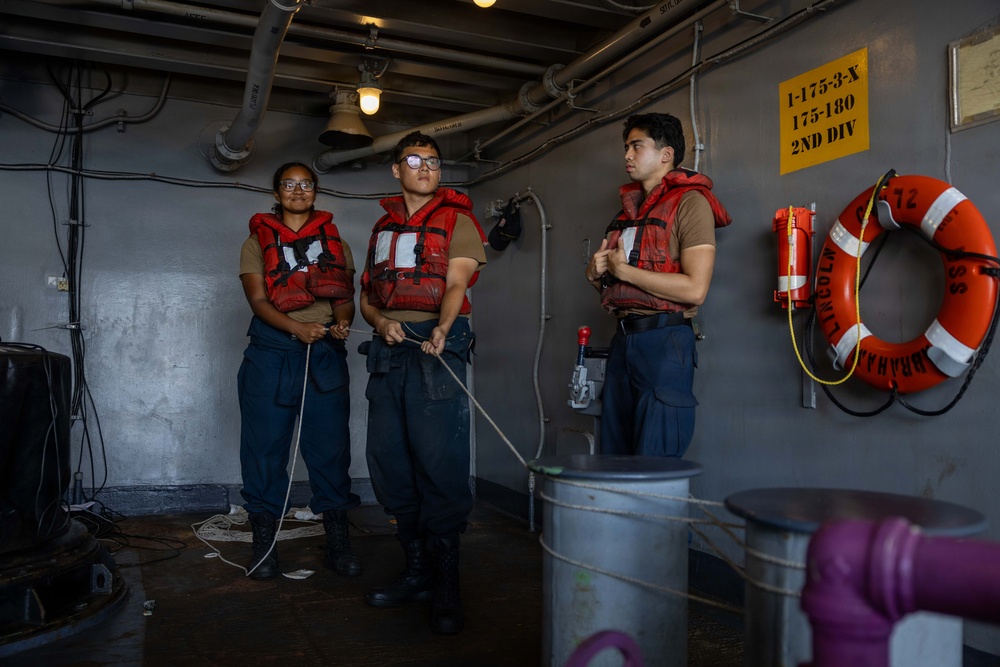 Three military personnel wearing red life vests are standing in what appears to be a naval vessel's interior deck area. Two of the individuals are holding rope lines, while a third appears to be observing or receiving instruction. Safety equipment including a life ring labeled 'Abraham Lincoln' and fire suppression equipment are mounted on the gray bulkhead wall.